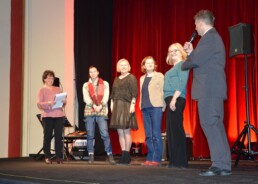 Cztery kobiety stoją w rzędzie na scenie. Piąta kobieta stoi po lewej stronie z kartkami papieru. Mężczyzna stoi po prawej stronie z mikrofonem w ręku.