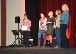 Cztery kobiety stoją w rzędzie na scenie. Po ich lewej stronie piąta kobieta trzyma mikrofon i kartki papieru. Patrzy w stronę publiczności.